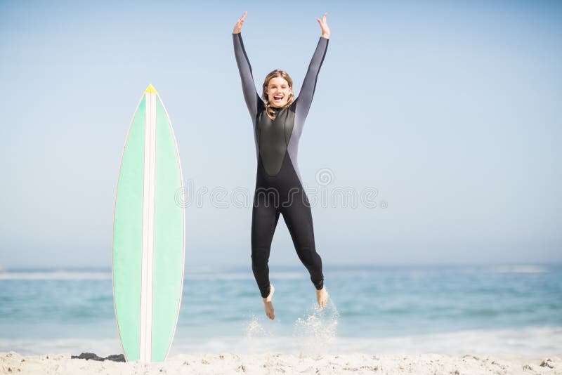 Femme enthousiaste dans le wetsuit sautant à côté de sa planche de surf photographie stock Femme enthousiaste dans le wetsuit sautant à côté de sa planche de surf photographie stock