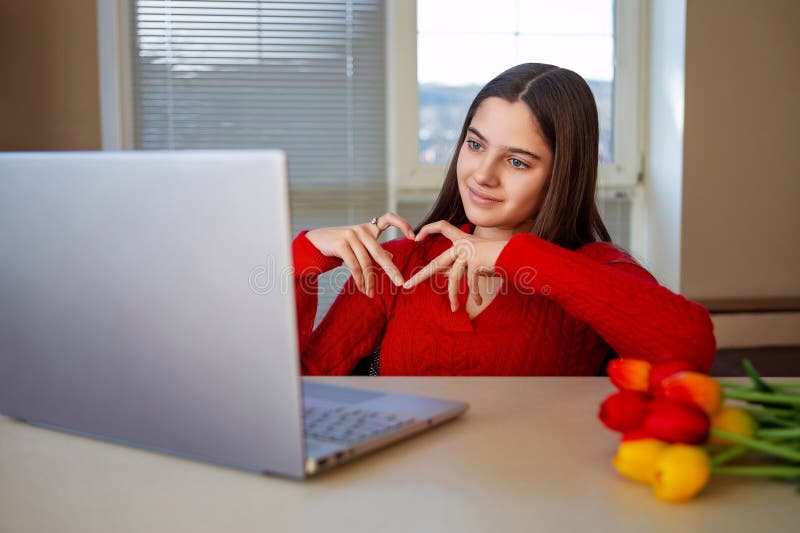 Femme faisant un geste cardiaque pendant un appel vidéo. Une jeune femme en chandail rouge fait un geste cardiaque avec ses mains photo stock
