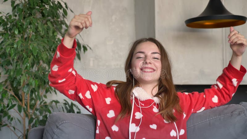 Femme heureuse de gingembre dans des pyjamas dansant et musique de écoute par des écouteurs, se reposant sur le divan dans le sal banque de vidéos