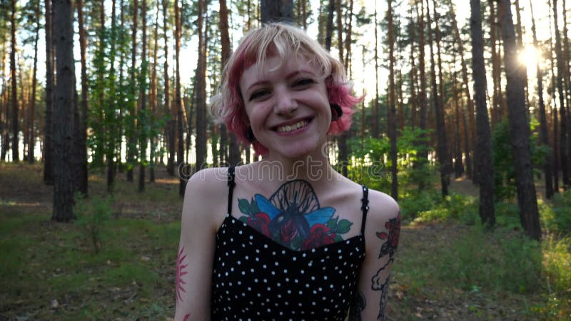 Femme hippie insouciante avec tatouages sur le chemin de la forêt. fille punk souriante heureuse avec cheveux roses regardant vers clips vidéos