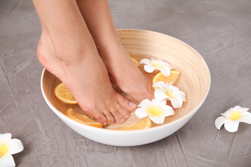 Femme imbibant de ses pieds dans la cuvette l'eau, les tranches oranges et les fleurs sur le fond gris, plan rapproché photos stock