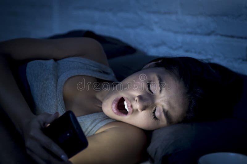 Femme latine sur le lit tard au service de mini-messages de nuit utilisant le baîllement de téléphone portable somnolent et fatig photographie stock Femme latine sur le lit tard au service de mini-messages de nuit utilisant le baîllement de téléphone portable somnolent et fatig photographie stock