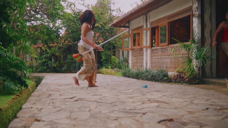 Femme marchant avec une arrosoir près d'une maison traditionnelle avec verdure luxuriante