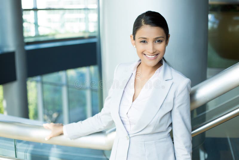 Closeup portrait, young professional, beautiful confident woman in gray white suit, friendly personality, holding rail, smiling indoors office background. Positive human emotions. Closeup portrait, young professional, beautiful confident woman in gray white suit, friendly personality, holding rail, smiling indoors office background. Positive human emotions