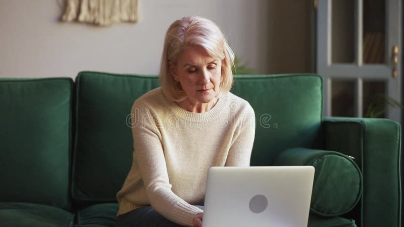 Femme mûre plus âgée sérieuse dactylographiant sur l'ordinateur portable se reposant sur le sofa banque de vidéos