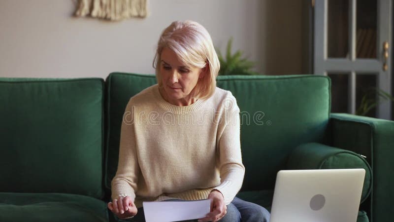 Femme mûre sérieuse calculant des documents de stock des effets utilisant l'ordinateur portable banque de vidéos
