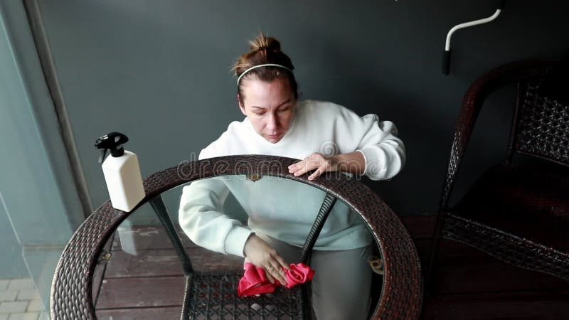 Femme nettoyant une table basse en verre sur un patio avec une bouteille de spray et un tissu pendant la journée banque de vidéos