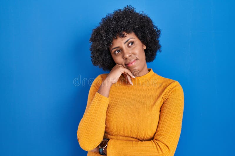 Femme noire aux cheveux bouclés debout sur fond bleu avec main sur le menton réfléchir à la question expression pensive. sourire avec un visage réfléchi. concept de doute