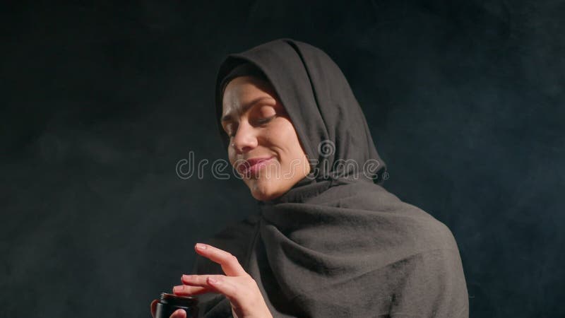 Femme portant le hijab noir applique doucement le produit de soin sa peau. Un éclairage doux crée une atmosphère calme la mettant banque de vidéos