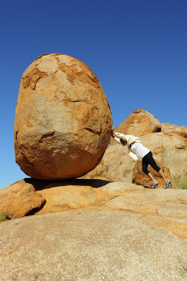 In a sisyphean task a woman in high heel shoes tries to push a huge boulder. In a sisyphean task a woman in high heel shoes tries to push a huge boulder.