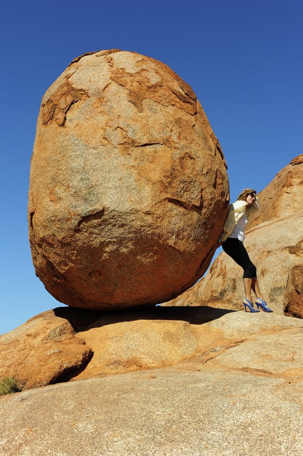 In a sisyphean task a woman in high heel shoes tries to push a huge boulder. In a sisyphean task a woman in high heel shoes tries to push a huge boulder.