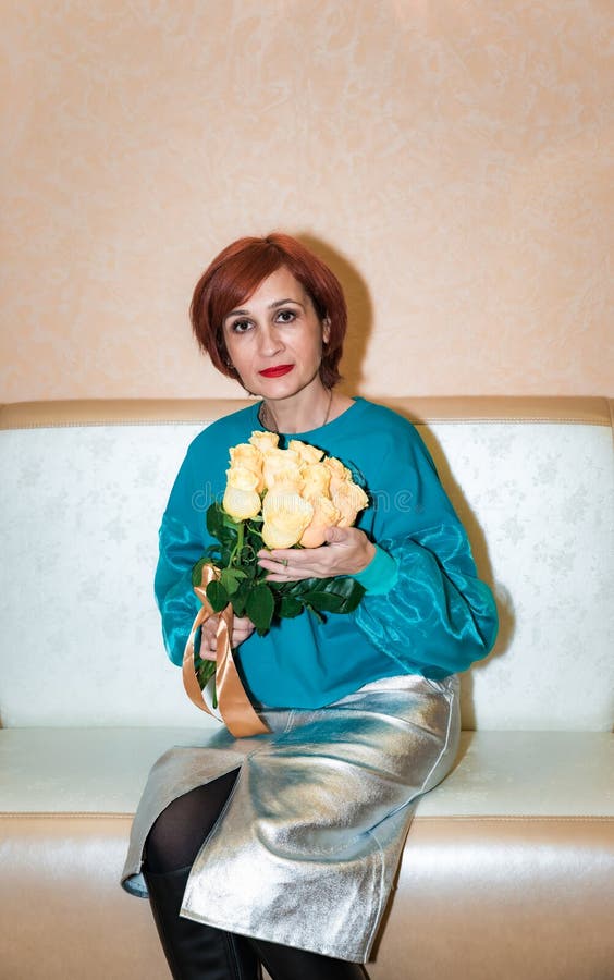 Femme russe de 48 ans assise à l'intérieur et tenant un bouquet de roses jaunes avec un ruban portant un blouse turquoise. photos stock
