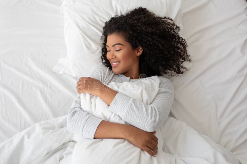 Hispanic woman with curly hair smiles contentedly as she embraces a fluffy pillow, lying in comfort on a bright white bedspread. Hispanic woman with curly hair smiles contentedly as she embraces a fluffy pillow, lying in comfort on a bright white bedspread