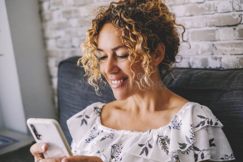 Femme souriant à l'intérieur à la maison et utilisant un téléphone mobile pour discuter et lire la notification ou surfer sur le net. femelle moderne assise sur canapé et écrivant sur appareil cellulaire avec application et gadgets. communication