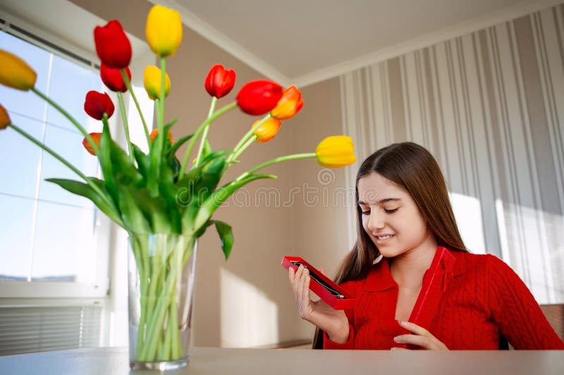 Femme souriante ouvrant le collier cadeau près des tulipes. Une jeune femme en chandail rouge sourit en ouvrant une boîte cadeau r photographie stock libre de droits