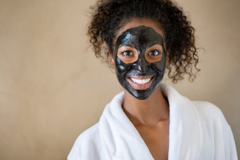 Smiling young woman with charcoal face mud looking at camera  on beige background with copy space. Portrait of african woman with curly hair wearing white bath robe getting black purifying mask at spa. Happy girl feeling relaxed at salon after face beauty facial treatment with peeling scrub clay. Smiling young woman with charcoal face mud looking at camera  on beige background with copy space. Portrait of african woman with curly hair wearing white bath robe getting black purifying mask at spa. Happy girl feeling relaxed at salon after face beauty facial treatment with peeling scrub clay