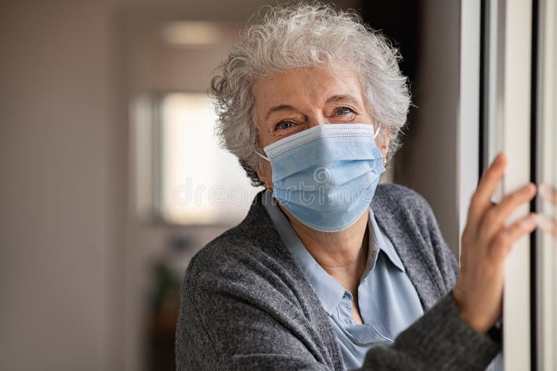 Portrait of smiling senior woman wearing face mask and standing near window. Old woman with grey hair wearing surgical mask and looking at camera during the lockdown. Happy elderly lady during the covid-19 pandemic stay at home. Portrait of smiling senior woman wearing face mask and standing near window. Old woman with grey hair wearing surgical mask and looking at camera during the lockdown. Happy elderly lady during the covid-19 pandemic stay at home