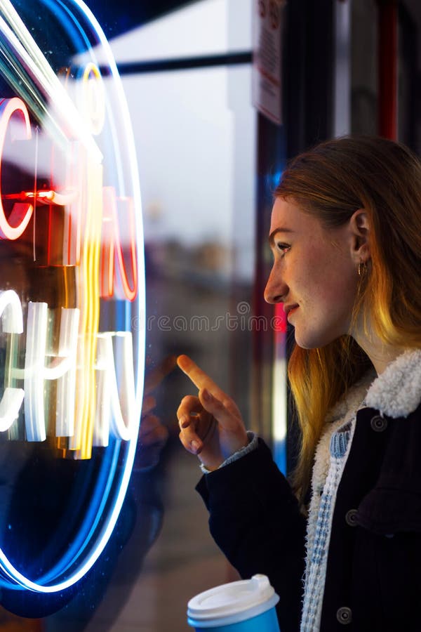 Young woman tourist with light makeup looks around standing near bar with colorful neon sign against night megalopolis. Young woman tourist with light makeup looks around standing near bar with colorful neon sign against night megalopolis.