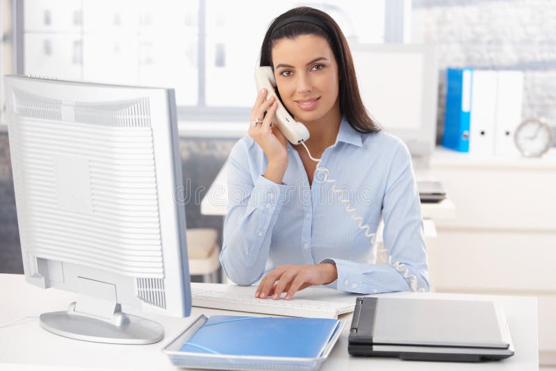 Portrait of smiling woman working in office, using computer and landline phone. Portrait of smiling woman working in office, using computer and landline phone.