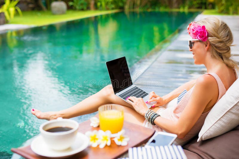 Femme travaillant sur l'ordinateur portable tout en se reposant par la piscine images stock Femme travaillant sur l'ordinateur portable tout en se reposant par la piscine images stock