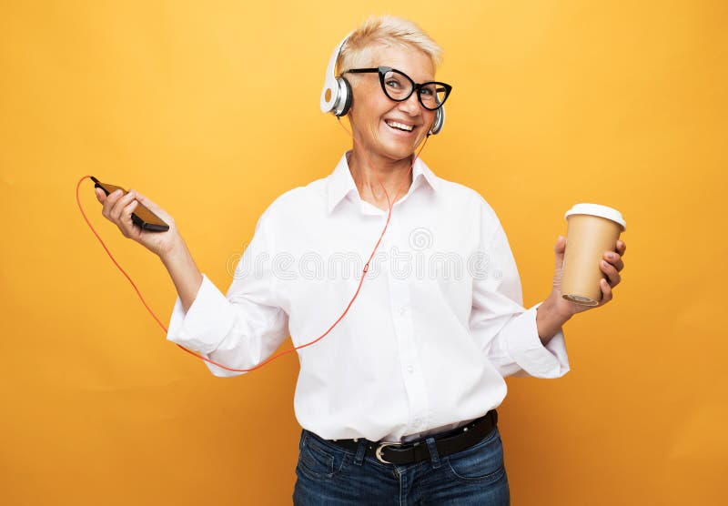 Femme âgée aux cheveux courts écoutant de la musique avec un casque et tenant du café pour passer sur fond jaune. image libre de droits