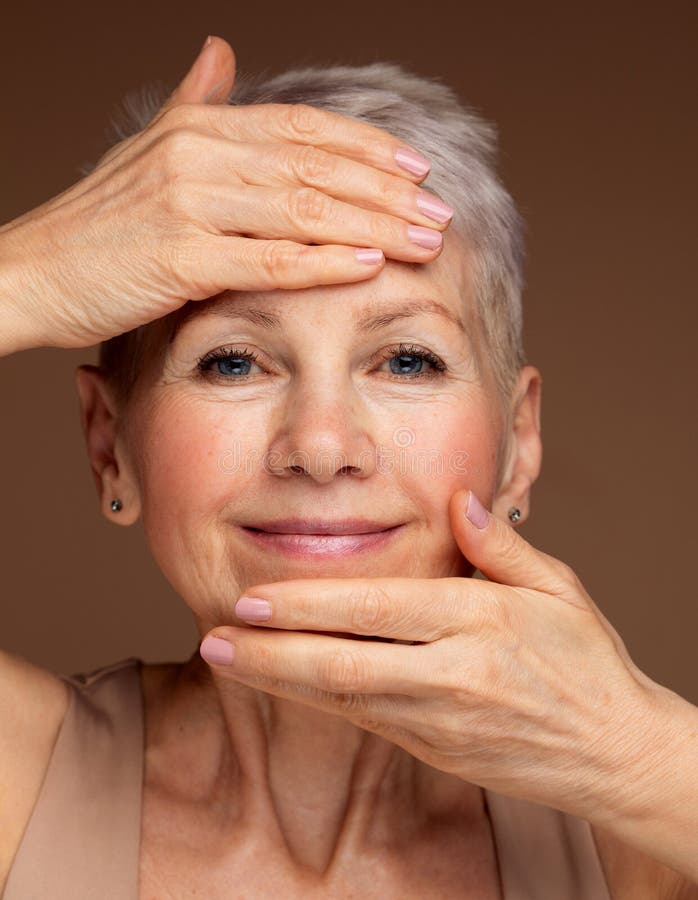 Femme âgée âgée de 60 ans qui regarde l'appareil photo qui touche son visage sur fond brun. photographie stock libre de droits