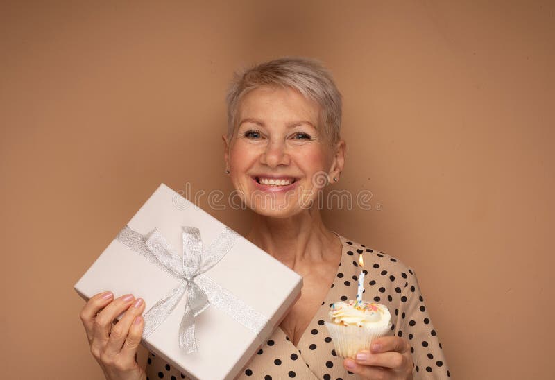 Femme âgée gaie avec une coupe de cheveux courte tient un cupcake avec une bougie et présente boîte fête un anniversaire. photo stock