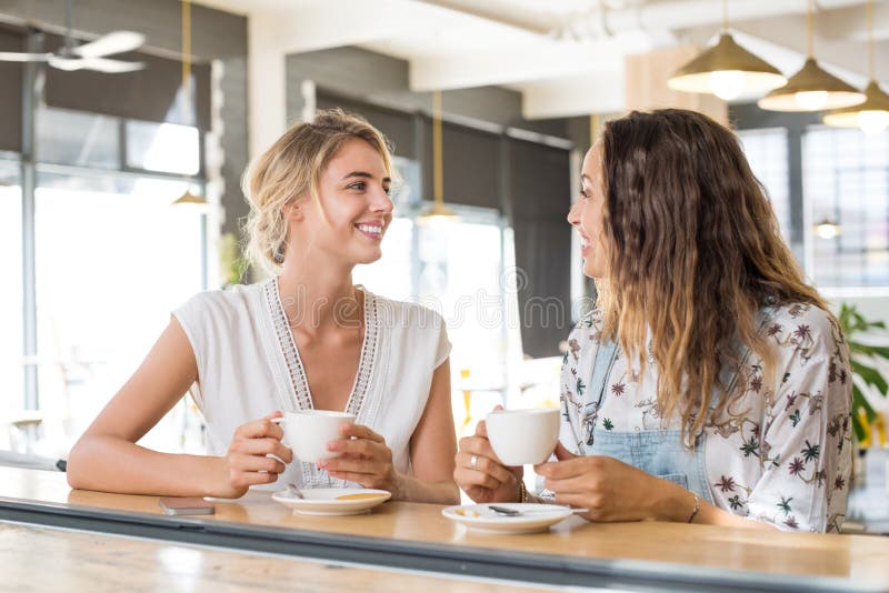 Two beautiful young women holding cup of coffee and talking to each other. Young blonde women in conversation with her best friend while sipping a cup of capuccino. Friends meeting up for coffee after a long time. Two beautiful young women holding cup of coffee and talking to each other. Young blonde women in conversation with her best friend while sipping a cup of capuccino. Friends meeting up for coffee after a long time.