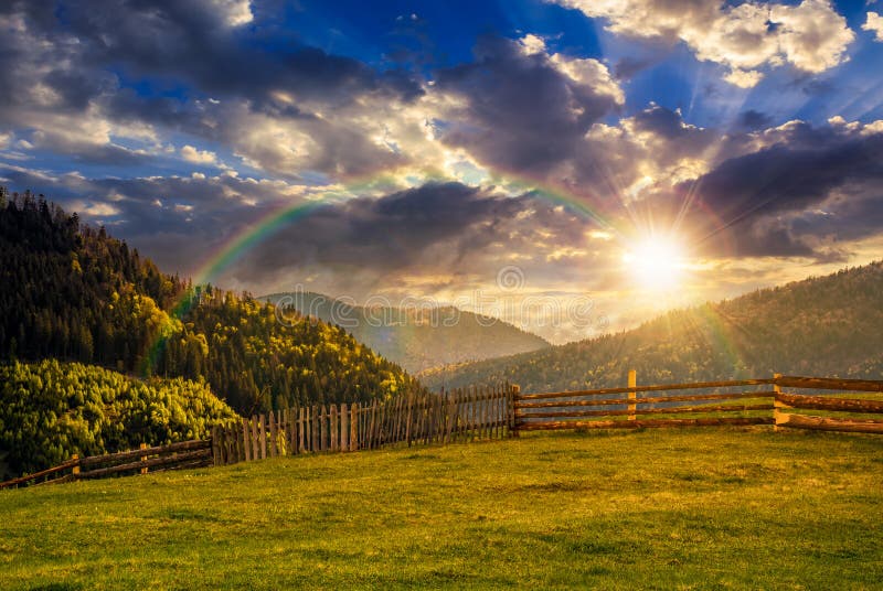 Fence through the grassy meadow in mountains at sunset with rain