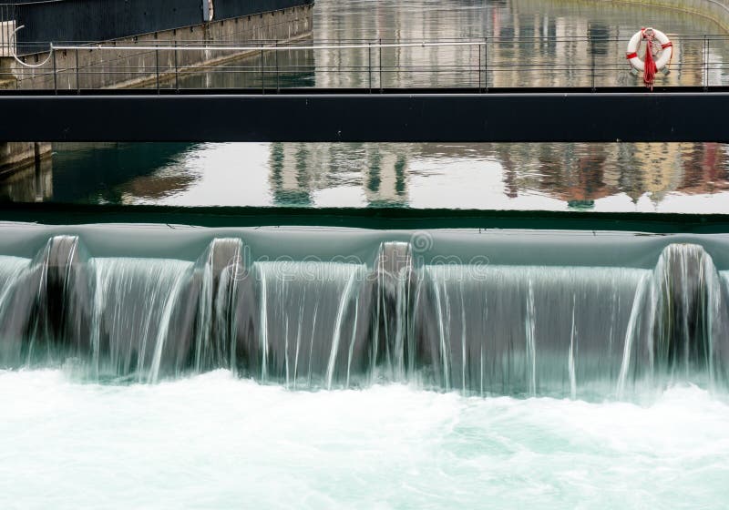 Fermez-vous d'un déversoir concret synthétique dans une ville avec une rivière bleue cascadant au-dessus de elle et une passerell photo stock Fermez-vous d'un déversoir concret synthétique dans une ville avec une rivière bleue cascadant au-dessus de elle et une passerell photo stock