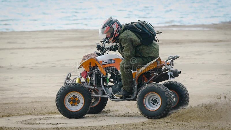 Fermez-vous de l'homme dans l'équipement de protection et du casque dérivant sur son ATV sur la plage avec la mer à l'arrière-pla