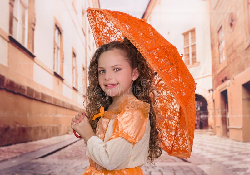 Fermez-vous de la petite belle fille bouclée utilisant un beau costume colonial et jugeant un parapluie orange dans brouillé image libre de droits Fermez-vous de la petite belle fille bouclée utilisant un beau costume colonial et jugeant un parapluie orange dans brouillé image libre de droits