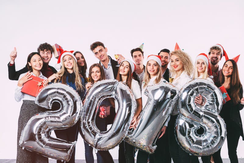 Office christmas party 2018. Group of cheerful young people in Santa hats holding silver colored number balloons. Celebration concept. Office christmas party 2018. Group of cheerful young people in Santa hats holding silver colored number balloons. Celebration concept.