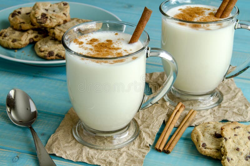 Glass mugs filled with homemade egg nog with cinnamon sticks and chocolate chip cookies on blue wooden table. Glass mugs filled with homemade egg nog with cinnamon sticks and chocolate chip cookies on blue wooden table