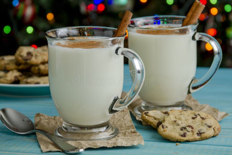 Glass mugs filled with homemade egg nog with cinnamon sticks and chocolate chip cookies on blue wooden table and Christmas tree lights in background. Glass mugs filled with homemade egg nog with cinnamon sticks and chocolate chip cookies on blue wooden table and Christmas tree lights in background