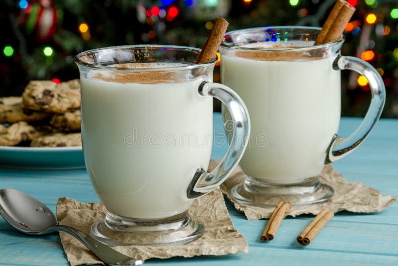 Glass mugs filled with homemade egg nog with cinnamon sticks and chocolate chip cookies on blue wooden table and Christmas tree lights in background. Glass mugs filled with homemade egg nog with cinnamon sticks and chocolate chip cookies on blue wooden table and Christmas tree lights in background