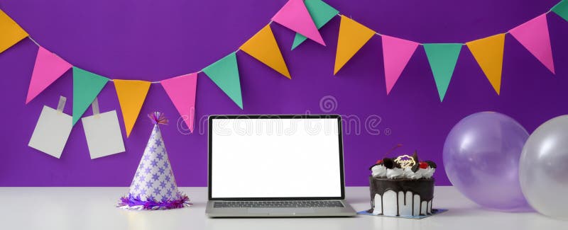 Festività online con cappello e palloncino per torte in un portatile a schermo bianco. Festival online di compleanno con schermo bianco per la torta al computer portatile con cappello e palloncino su tavolo bianco con decorazioni su muro viola