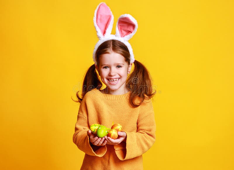 Fetiță amuzantă și fericită cu ouă de Paști și urechi de iepuraș pe galben stock images