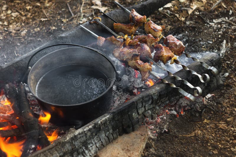 Field kitchen tourist camp on open fire of the bonfire, boiling water in boiler and grilled pork meat skewers royalty free stock photo Field kitchen tourist camp on open fire of the bonfire, boiling water in boiler and grilled pork meat skewers royalty free stock photo