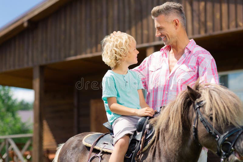 Figlio bionda-dai capelli riccio che si siede sul cavallo e che parla con papà immagine stock Figlio bionda-dai capelli riccio che si siede sul cavallo e che parla con papà immagine stock