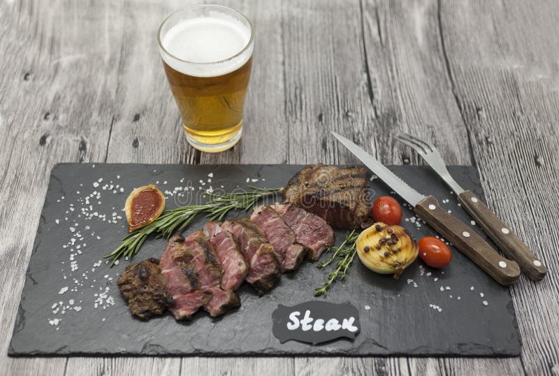 Filete asado a la parrilla del striploin en una placa de piedra con un vidrio de cerveza con una bifurcación y un cuchillo fotos de archivo Filete asado a la parrilla del striploin en una placa de piedra con un vidrio de cerveza con una bifurcación y un cuchillo fotos de archivo