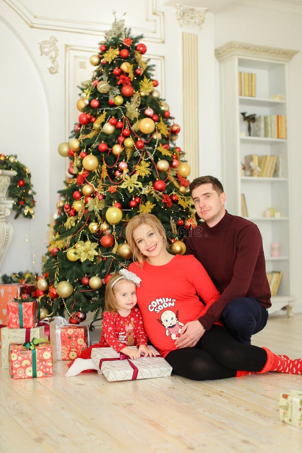 Filha agradável pequena que senta-se com pai e a mãe grávida perto da árvore de Natal e que mantém presentes foto de stock Filha agradável pequena que senta-se com pai e a mãe grávida perto da árvore de Natal e que mantém presentes foto de stock