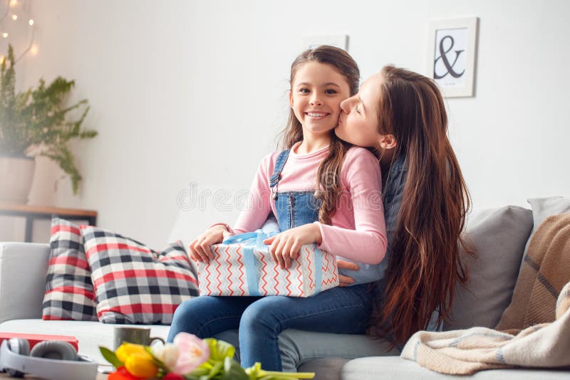 Filha de assento do aniversário da mãe e da filha em casa que mantém a caixa atual beijada pela mamã imagem de stock royalty free Filha de assento do aniversário da mãe e da filha em casa que mantém a caixa atual beijada pela mamã imagem de stock royalty free