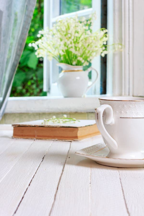 A Cup of tea or coffee and a book on a white wooden vintage retro table and a bouquet of Lily of the valley flowers on the windowsill by the open window in the rustic house in the spring morning. A Cup of tea or coffee and a book on a white wooden vintage retro table and a bouquet of Lily of the valley flowers on the windowsill by the open window in the rustic house in the spring morning