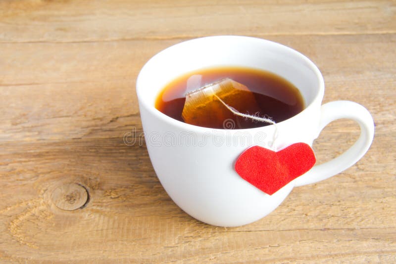 White cup of tea with red heart shaped label on teabag on wooden background (surface). Love and care concept. White cup of tea with red heart shaped label on teabag on wooden background (surface). Love and care concept.