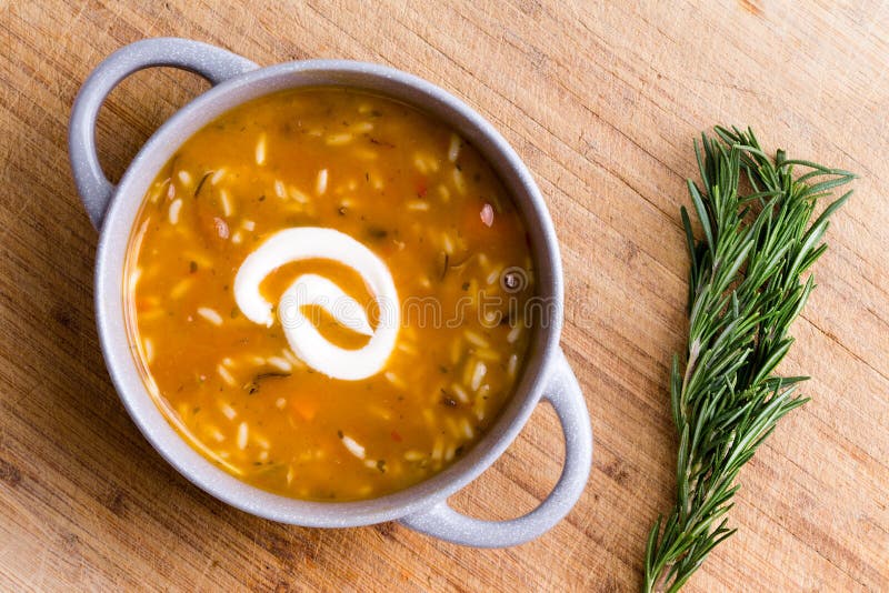 Cup of chicken broth with wild rice served with a twirl of sour cream ready to be garnished with fresh rosemary displayed alongside on a bamboo cutting board, overhead view. Cup of chicken broth with wild rice served with a twirl of sour cream ready to be garnished with fresh rosemary displayed alongside on a bamboo cutting board, overhead view