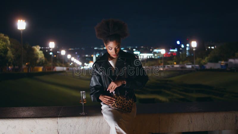 Fille aux cheveux tenant du verre de vin assis aux lumières de la rue de nuit. femme africaine banque de vidéos