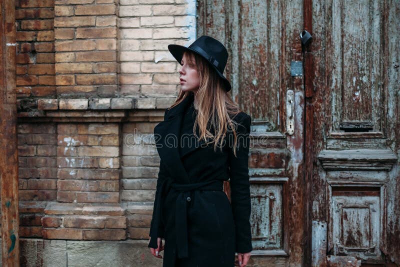 Fille blonde avec de longs cheveux, dans le manteau noir dans le chapeau, supports sur le fond maison de brique de vieille porte photo libre de droits Fille blonde avec de longs cheveux, dans le manteau noir dans le chapeau, supports sur le fond maison de brique de vieille porte photo libre de droits