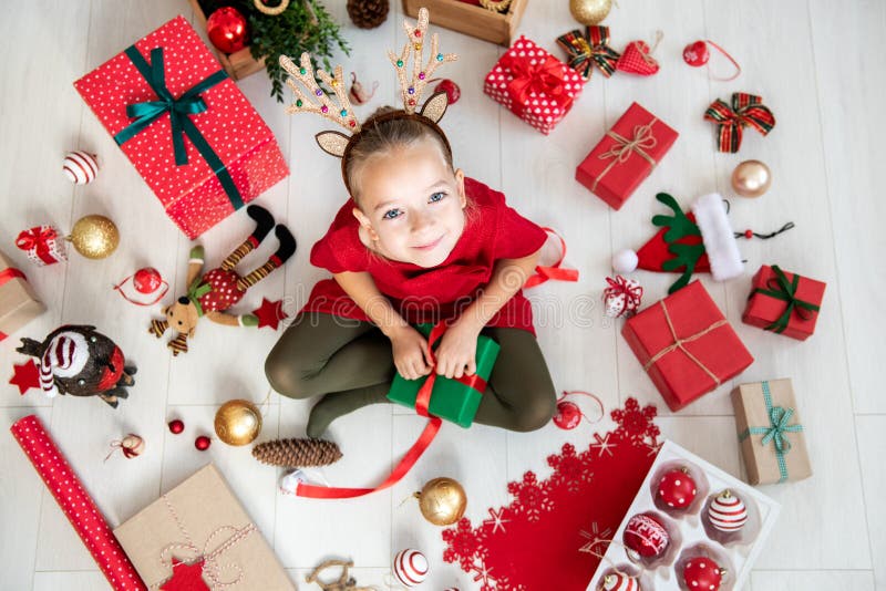Fille curieuse utilisant des andouillers de renne de costume de Noël se reposant sur le plancher, cadeau de Noël s'ouvrant, vue s photographie stock libre de droits Fille curieuse utilisant des andouillers de renne de costume de Noël se reposant sur le plancher, cadeau de Noël s'ouvrant, vue s photographie stock libre de droits
