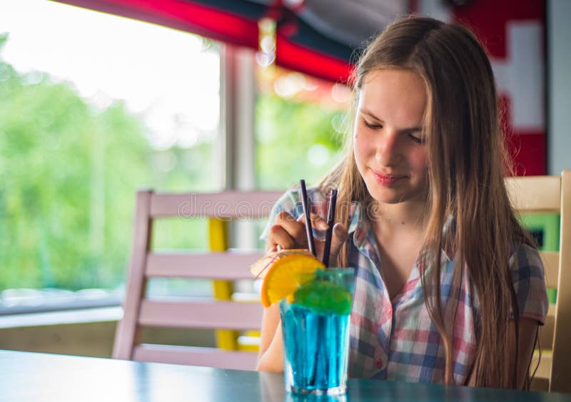Fille de brune d'adolescent avec de longs cheveux tenant un verre avec un cocktail bleu de limonade avec des fruits à une table e image stock Fille de brune d'adolescent avec de longs cheveux tenant un verre avec un cocktail bleu de limonade avec des fruits à une table e image stock